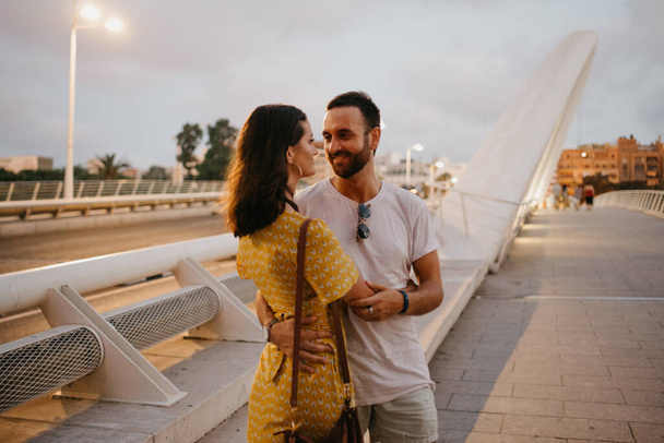 Una chica morena con un vestido amarillo y su novio sonriente se abrazan en un puente blanco en Valencia. Un par de turistas en una cita en la cálida noche. - Foto, imagen