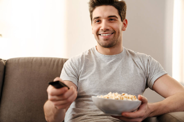 Happy man watching tv and eating popcorn at home on a couch - Fotografie, Obrázek