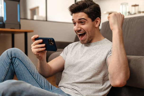 Delighted guy making winner gesture while playing video game on cellphone at home - Photo, Image