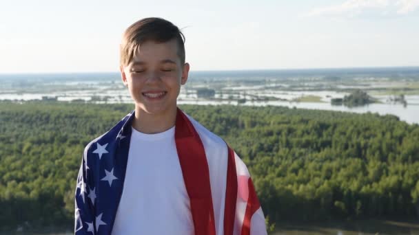 Ragazzo biondo sventola bandiera nazionale USA all'aperto sopra il cielo blu sulla riva del fiume - Filmati, video