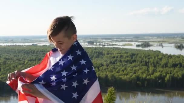 Niño rubio ondeando bandera nacional de EE.UU. al aire libre sobre el cielo azul en la orilla del río - Metraje, vídeo