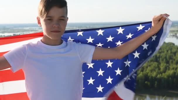 Menino loiro acenando bandeira nacional dos EUA ao ar livre sobre o céu azul na margem do rio - Filmagem, Vídeo