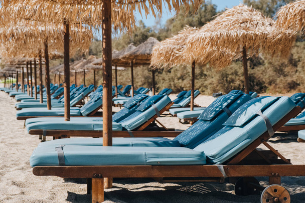 ギリシャのミコノス島のビーチで傘の下に空の青い太陽のベッドとラウンジャーの行のサイドビュー. - 写真・画像