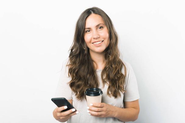 Retrato de mujer joven sonriente sosteniendo teléfono inteligente y taza de café de papel. - Foto, imagen