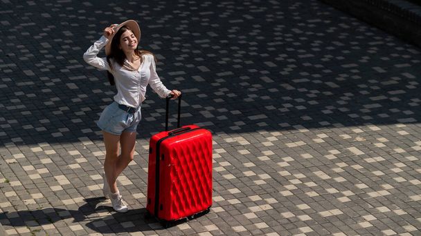 Happy caucasian young woman in hat and shorts holding a large red suitcase outdoors. - Fotografie, Obrázek