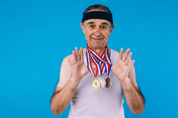 Elderly athlete with sun marks on his arms, with three medals on his neck, teaching them, on blue background - Photo, Image