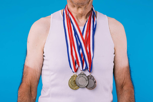 Ηλικιωμένος αθλητής με σημάδια από τον ήλιο στα χέρια του, με τρία μετάλλια στο λαιμό του για ένα αθλητικό επίτευγμα σε μπλε φόντο - Φωτογραφία, εικόνα