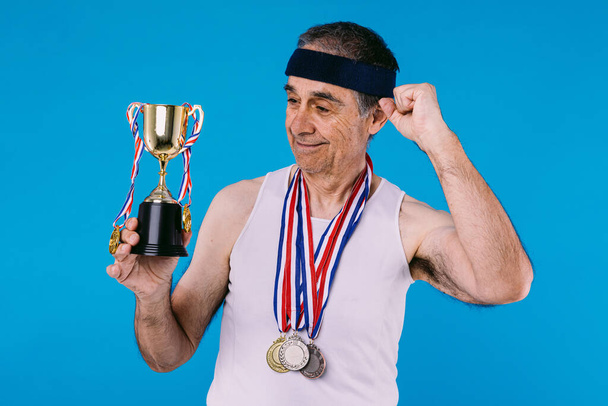 Elderly athlete with sun marks on his arms, with three medals on his neck and a trophy in his hands, clenching his fist as a sign of strength, on blue background - Photo, Image