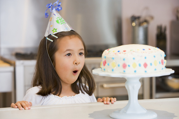 Young girl wearing party hat at kitchen counter looking at cake - Фото, изображение