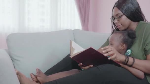 Jonge Afrikaanse moeder en dochter doen activiteit met het vertellen van verhalen op de bank samen thuis, moeder lezen verhaal tijd met een klein kind op de bank in de woonkamer met gezellige, familie en onderwijs concept. - Video