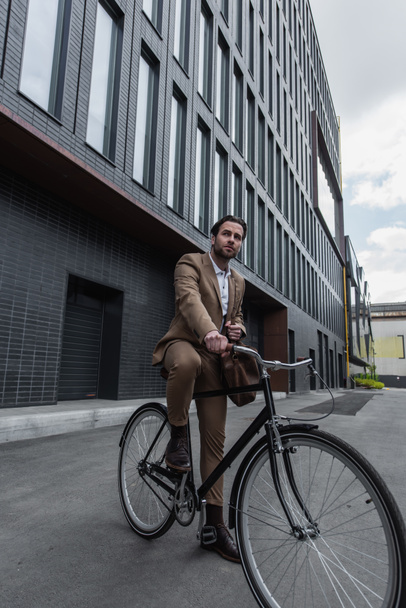 full length of young businessman in suit with leather bag riding bicycle outside  - 写真・画像