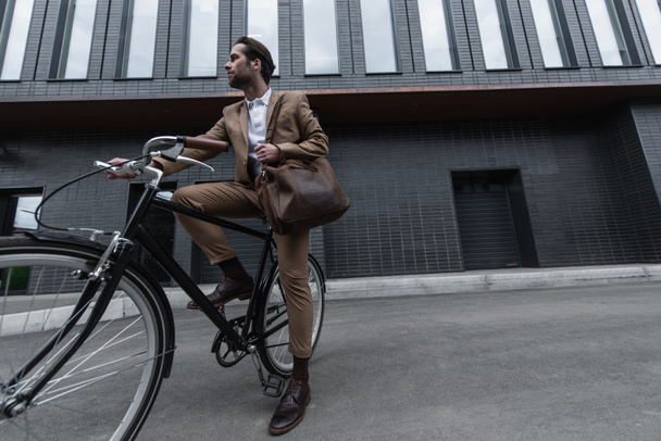 longitud completa de joven hombre de negocios en ropa formal con bolsa de cuero montar en bicicleta fuera  - Foto, Imagen
