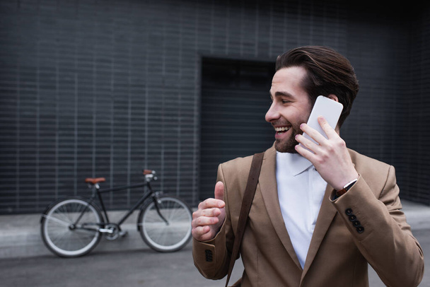 happy young businessman in formal wear talking on cellphone near building  - Фото, изображение