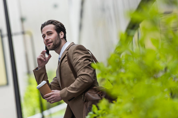 homme souriant en tenue formelle parlant sur téléphone portable et tenant tasse en papier près de plante floue  - Photo, image