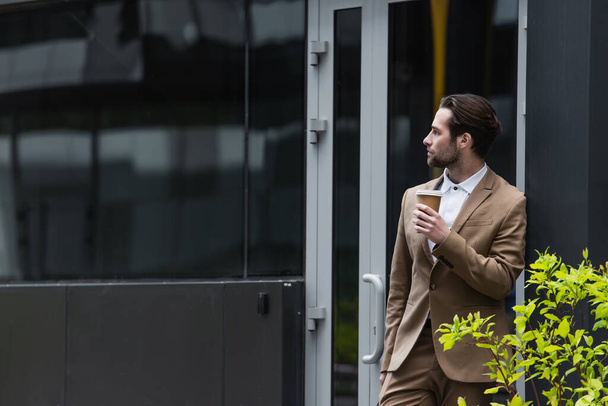thoughtful businessman in formal wear holding coffee to go near building - Foto, Imagen