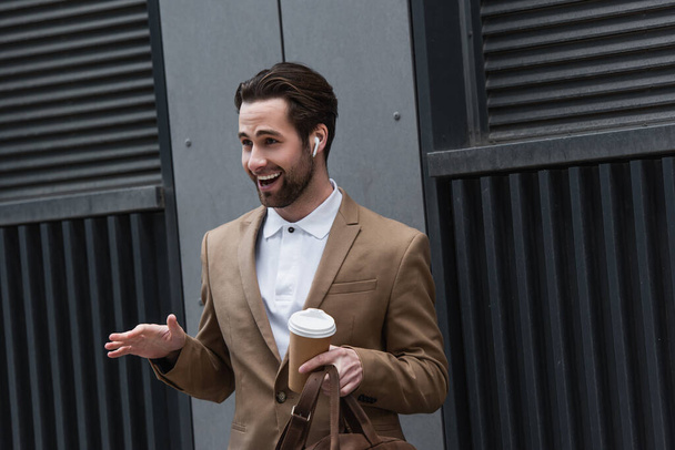 cheerful businessman in earphones holding coffee to go near building - Foto, afbeelding