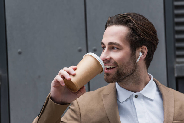 happy businessman in earphones drinking coffee to go near building - Photo, Image