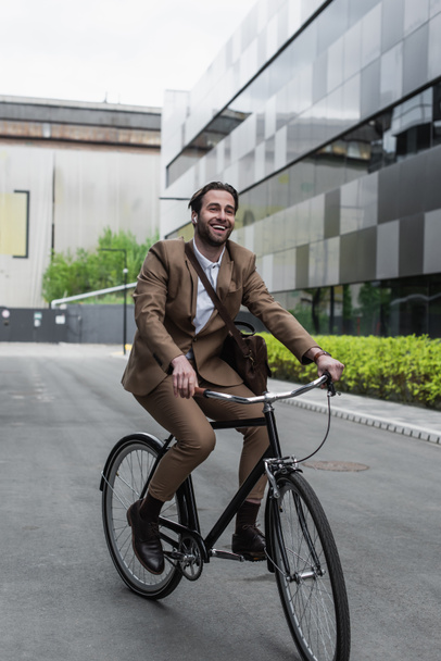 full length of cheerful businessman in earphones riding bicycle near building - Foto, Bild
