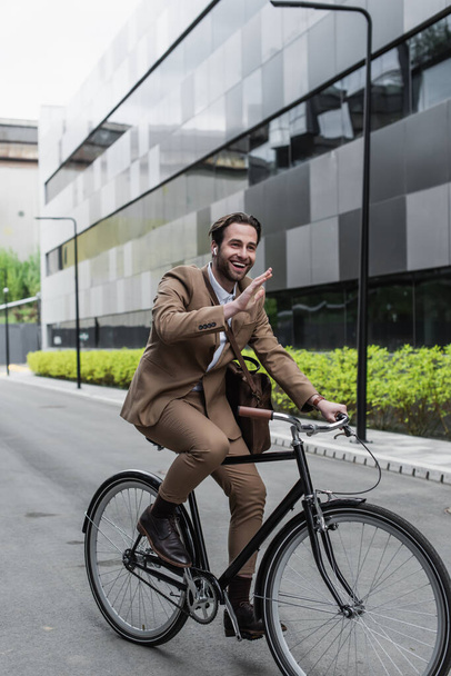 full length of cheerful businessman in earphones riding bicycle and waving hand near building and plants  - Foto, immagini