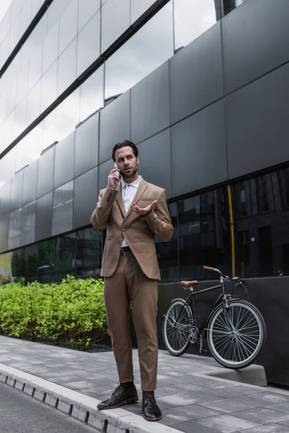 longitud completa de hombre de negocios en traje hablando en el teléfono celular cerca de la bicicleta y la construcción  - Foto, Imagen