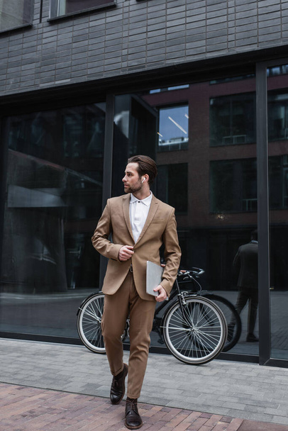 full length of happy businessman in earphones walking and holding laptop near building and bike  - 写真・画像