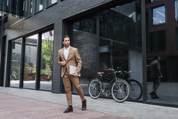 full length of happy businessman in earphones walking and holding laptop near building and bicycle  - 写真・画像