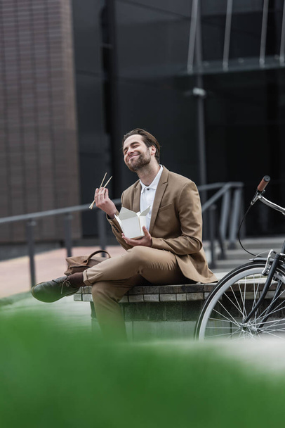 pleased businessman in suit holding cardboard box with asian food and chopsticks near bicycle outside  - Foto, Bild