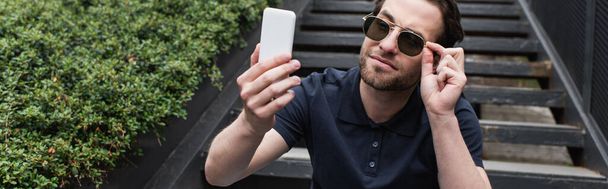 Güneş gözlüklü ve polo tişörtlü mutlu adam dışarıda cep telefonuyla selfie çekiyor. - Fotoğraf, Görsel