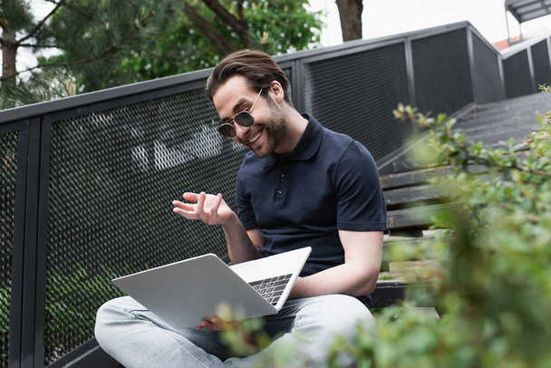 gelukkig man in zonnebril en polo shirt gebaren terwijl het hebben van video chat buiten  - Foto, afbeelding