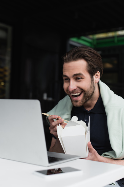 hombre feliz en polo y sudadera sosteniendo palillos y caja de cartón mientras mira el ordenador portátil cerca de teléfono inteligente borroso  - Foto, Imagen