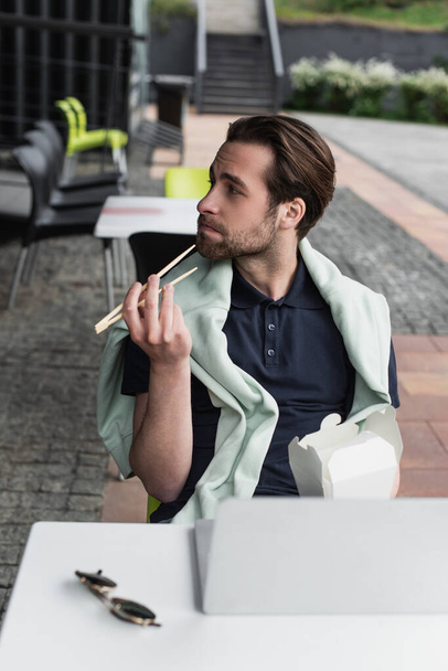 Polo tişörtlü sakallı adam elinde yemek çubukları ve karton kutuyla masada bulanık güneş gözlüğü takıyor. - Fotoğraf, Görsel