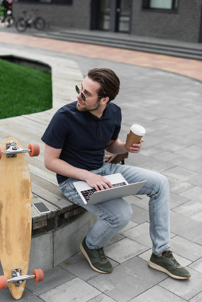 hombre en gafas de sol y polo sosteniendo la taza de papel y usando el ordenador portátil cerca del teléfono inteligente y longboard  - Foto, imagen
