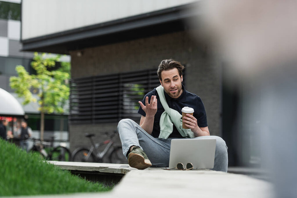 бородатый мужчина жестикулирует во время видеозвонка на ноутбуке, держа в руках бумажную чашку  - Фото, изображение