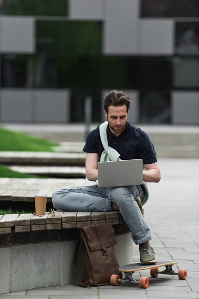 szakállas férfi pólóban és pulóverben laptoppal papír pohár és longboard közelében  - Fotó, kép