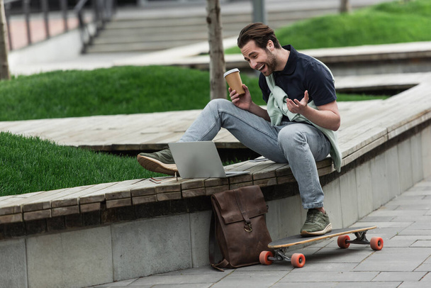 cheerful man in polo shirt and sweatshirt holding coffee to go and laughing while sitting near longboard and gadgets  - Photo, Image