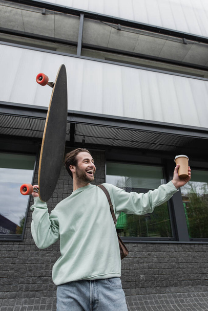 cheerful man in sweatshirt holding paper cup and longboard outside   - Foto, imagen