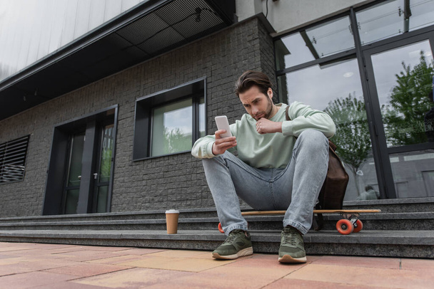 full length of man in sweatshirt looking at smartphone near paper cup and longboard outside   - Fotografie, Obrázek