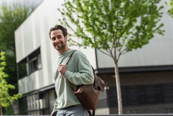 smiling bearded man in sweatshirt holding leather strap of backpack near building outside  - Fotó, kép