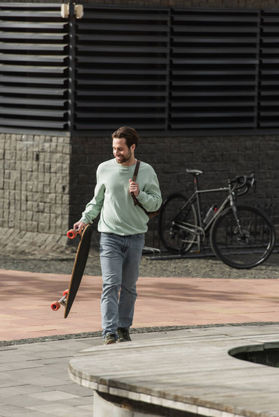 teljes hossza boldog szakállas férfi pulóverben gazdaság longboard és bőr heveder hátizsák séta közben kívül  - Fotó, kép