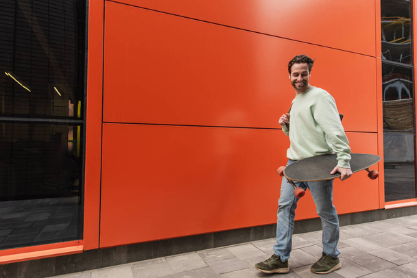 cheerful bearded man in sweatshirt holding longboard while walking near orange wall - Photo, image