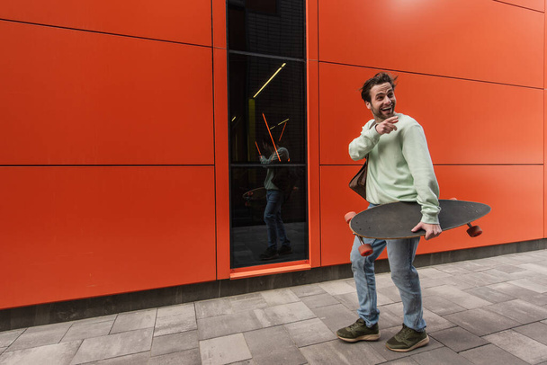 sonriente hombre con sudadera sosteniendo longboard mientras señala con el dedo cerca de la pared naranja - Foto, imagen