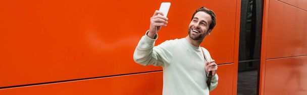 cheerful man in sweatshirt taking selfie on smartphone near orange wall, banner - Photo, image