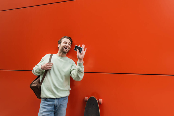vidám férfi pulóverben gazdaság okostelefon üres képernyő és integető kéz közel narancssárga fal  - Fotó, kép