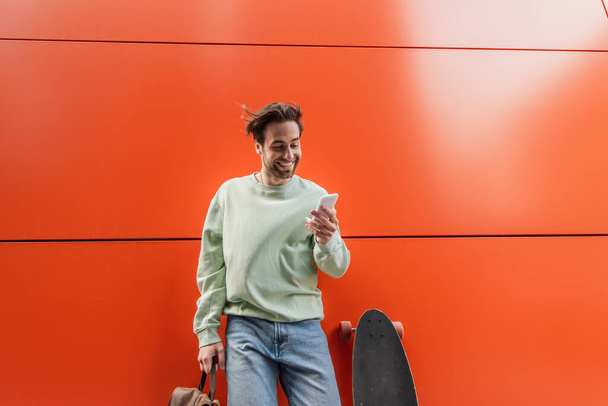 hombre alegre en sudadera que sostiene la mochila y el uso de teléfono inteligente cerca de longboard y pared naranja  - Foto, imagen
