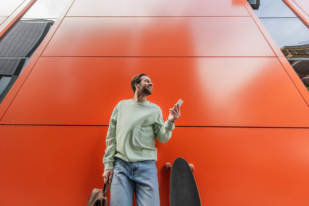 Tiefansicht eines fröhlichen Mannes im Sweatshirt mit Rucksack und Smartphone in der Nähe von Longboard und orangefarbener Wand  - Foto, Bild