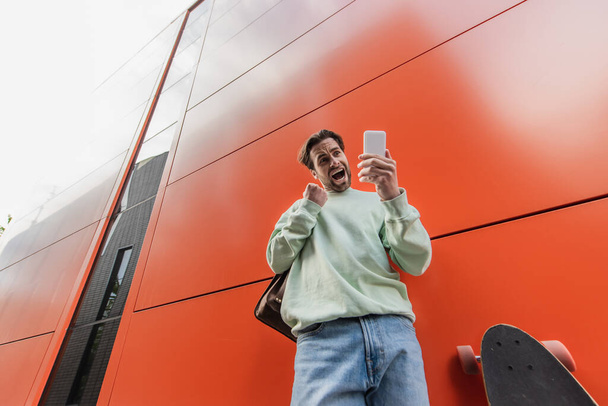 lage hoek van opgewonden man in sweatshirt met smartphone in de buurt van longboard en oranje muur  - Foto, afbeelding
