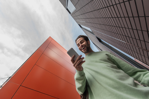 vista de ángulo bajo del hombre sorprendido en la sudadera que sostiene el teléfono inteligente cerca de edificios - Foto, imagen