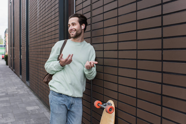 hombre feliz en sudadera y auriculares inalámbricos hablando y haciendo gestos fuera  - Foto, Imagen