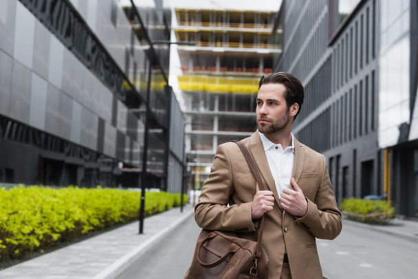 young businessman in suit holding leather strap of bag on urban street - Photo, image