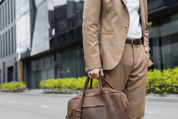 cropped άποψη του επιχειρηματία στο κοστούμι εκμετάλλευση δερμάτινη τσάντα και στέκεται με το χέρι στην τσέπη σε αστικό δρόμο - Φωτογραφία, εικόνα